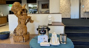 Ein Empfangsbereich mit einer Holzskulptur, einer Kaffeemaschine und einem Tisch mit Einwegbechern und einer Metallkanne.