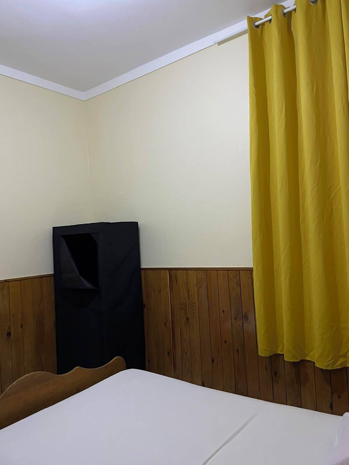 Une chambre d’hôtel avec un lit, une tête de lit en bois, un mur blanc et un rideau jaune.