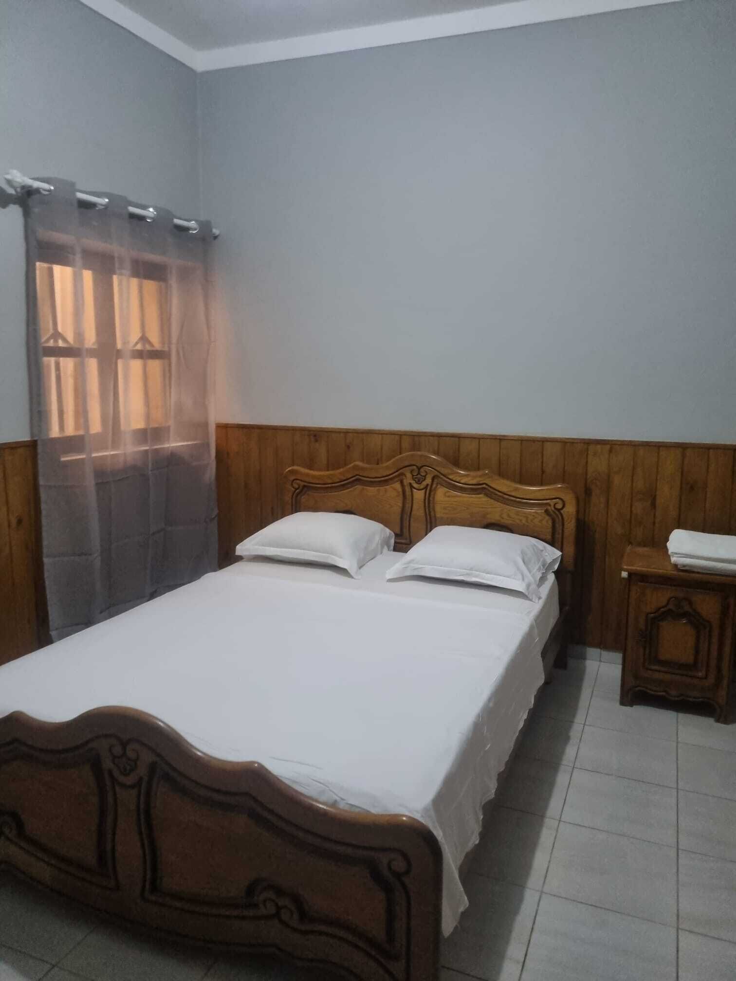A single bed with a wooden headboard, a small bedside table, and a window with a sheer curtain.