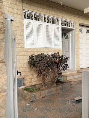 Una casa con fachada de piedra, contraventanas blancas, una puerta de garaje y un pequeño jardín con plantas.