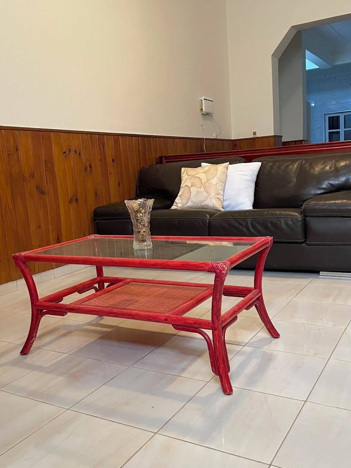 A living room with a glass-top red table, a black leather sofa with patterned cushions, and a wooden paneled wall.