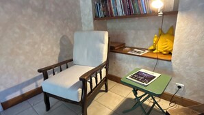 Una habitació amb una cadira de fusta, una taula petita amb una revista i una estanteria amb llibres i una làmpada.