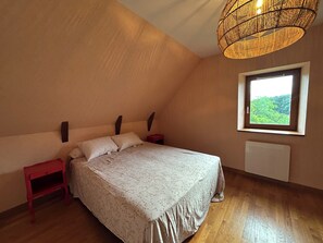 A bedroom with a sloped ceiling, a bed with a floral bedspread, a red nightstand, a window with a view of greenery, and a woven light fixture. - La Cerisaie - A house in the Nord/Aveyron region near Aubrac, Laguiole, and the Lot Valley. (Lassouts)