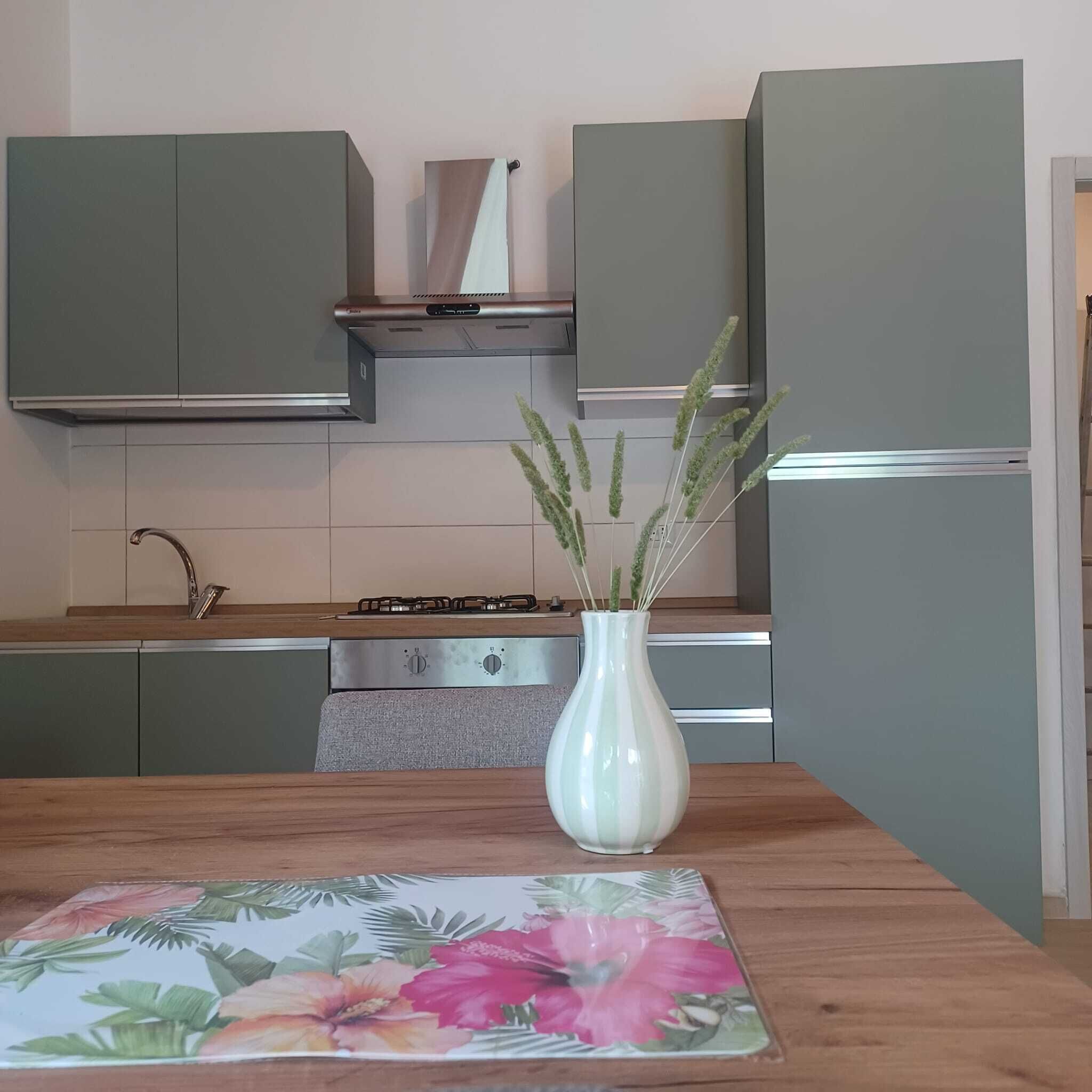 A kitchen with green cabinets, a white vase with plants, and a floral table mat.