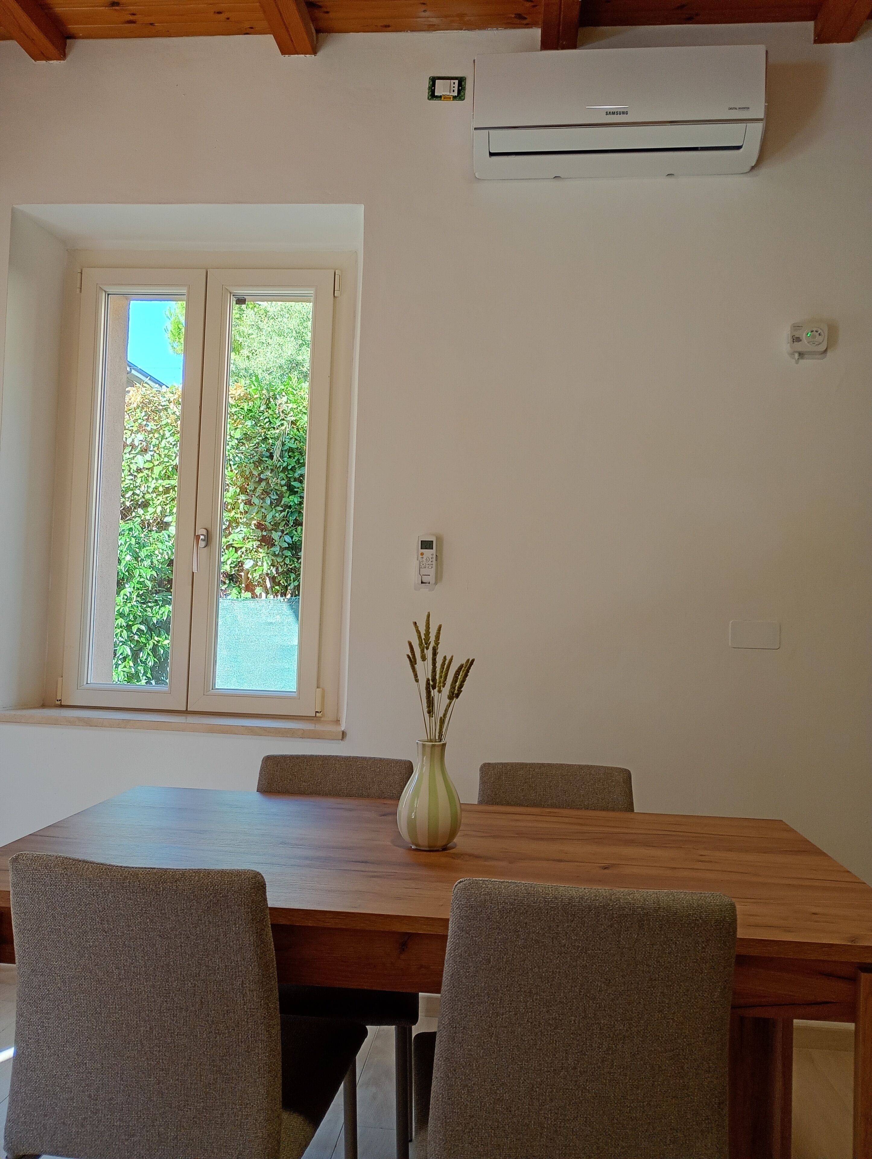 Un comedor con una mesa y sillas de madera, un jarrón con plantas secas, un equipo de aire acondicionado y una ventana con vistas al verdor.