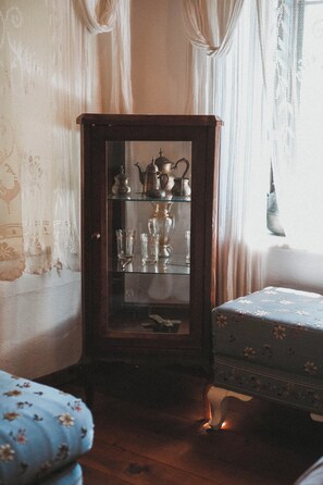 A wooden cabinet with glass doors containing tea sets and glasses, positioned next to a floral-patterned chair. - Boleka Bodrum (Bodrum)