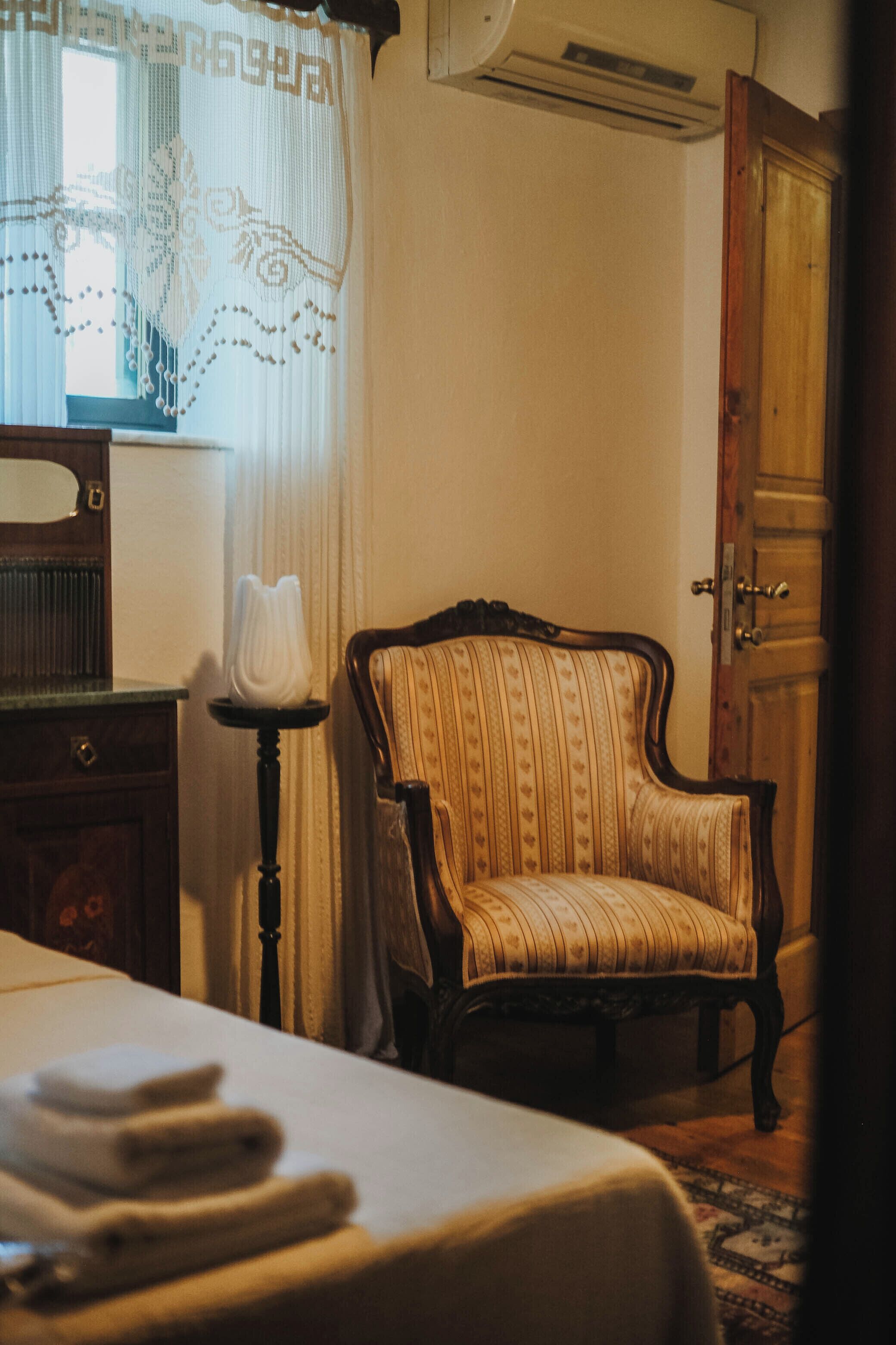 Una habitación con cama, una silla, lámpara, ventana con cortinas y un equipo de aire acondicionado.