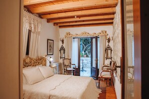 A bedroom with a wooden ceiling, a bed with a white bedspread, a chair, a table with a lamp, and a view of the outside through a glass door.