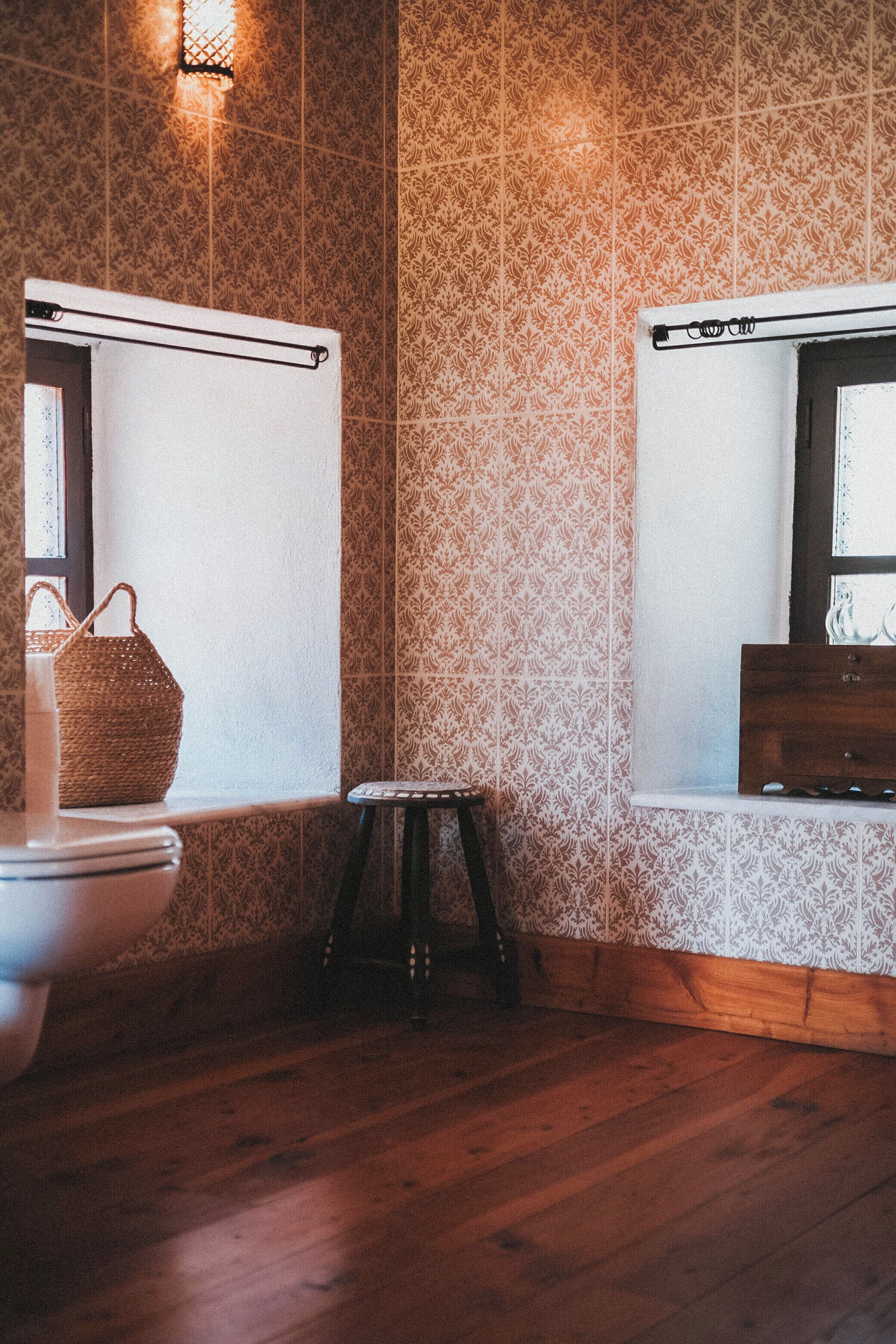 Une salle de bain avec un papier peint à motifs, un plancher en bois, un petit tabouret et un panier suspendu.