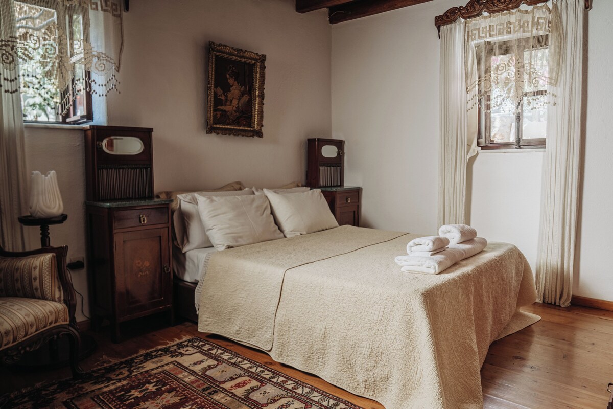 Un dormitorio con cama, mesita de noche, silla, ventana con cortinas y un cuadro en la pared.