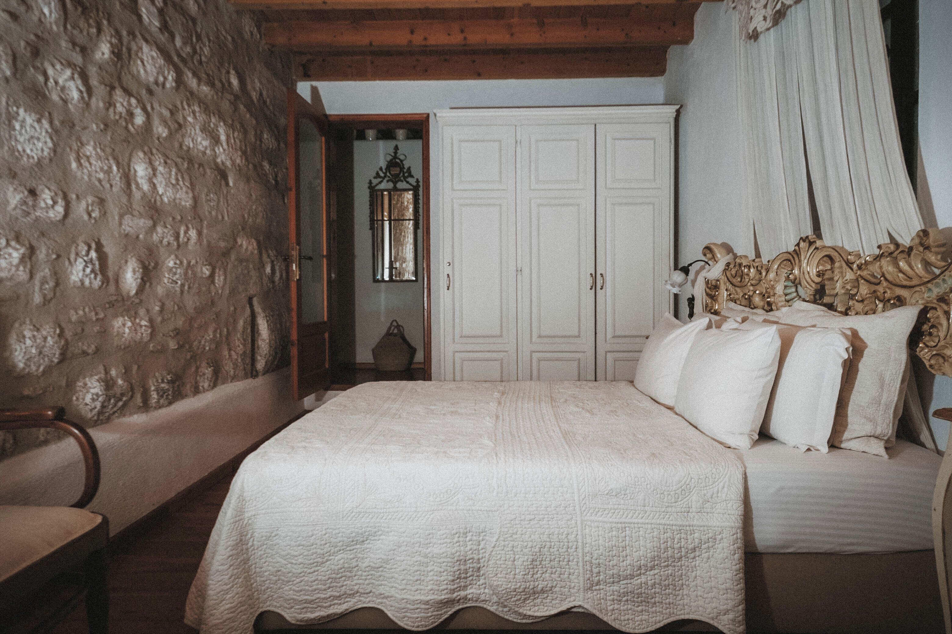 A bedroom with a stone wall, a wooden ceiling, a large bed with ornate headboard, and a white door.