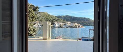 Vista a través de una puerta corredera de vidrio a un balcón con vista despejada al agua, un puente y un paisaje montañoso.