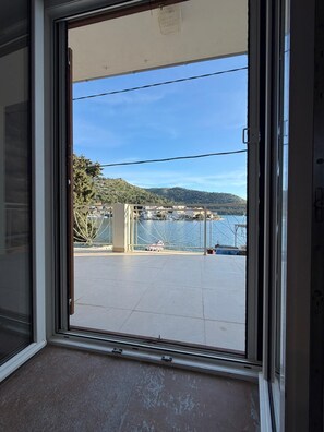 A view through a sliding glass door to a balcony with a clear view of a body of water, a bridge, and a hilly landscape. - Croatia, Dalmatia! Charming apartment in the fishing village of Blace, right on the water! (Blace)