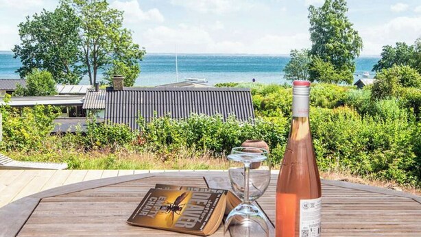 Ein Holztisch mit einer Weinflasche, einem Glas und einem Buch, vor einer Küstenlandschaft mit Grünflächen und einem Gebäude.