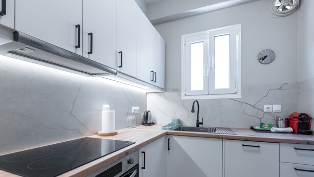 Una cocina moderna con armarios blancos, una placa de inducción negra, horno empotrado, ventana con persianas y una cafetera roja.