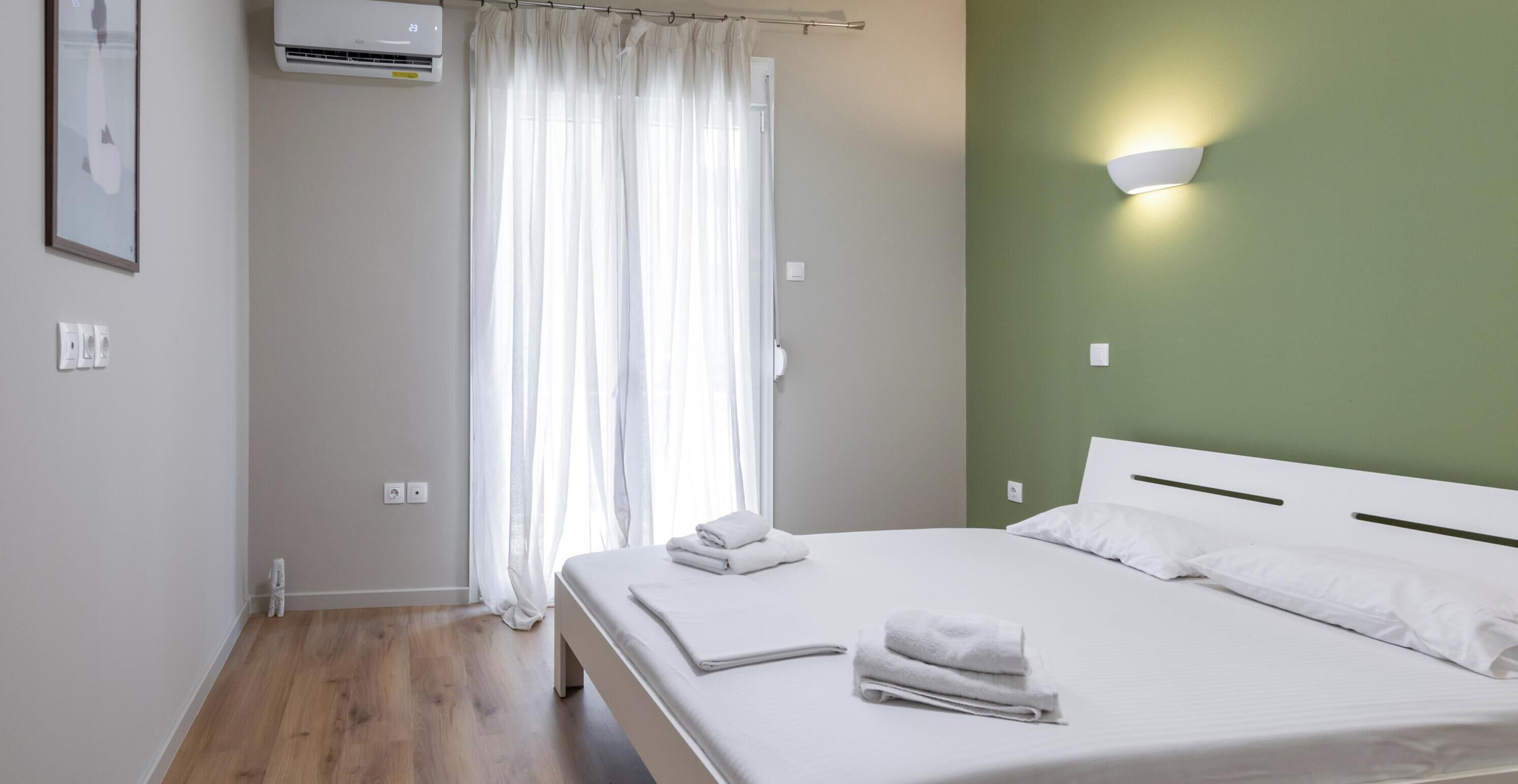A hotel room with a bed, white curtains, an air conditioning unit, and a wall-mounted light fixture.