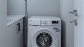 Lavadora blanca compacta con panel de control, ubicada en un lavadero pequeño que tiene una puerta y una escoba apoyada contra la pared.