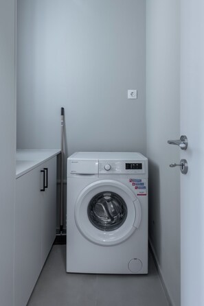 A compact, white washing machine with a control panel, located in a small laundry room with a door and a broom leaning against the wall. - Charming 2BR Downtown Metro (Athina)