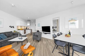 A modern living space with a sofa, ottoman, dining table, chairs, a TV, and a kitchen area. - Modern apartment with exceptional sea view and balcony at Marina Wendtorf (Wendtorf)