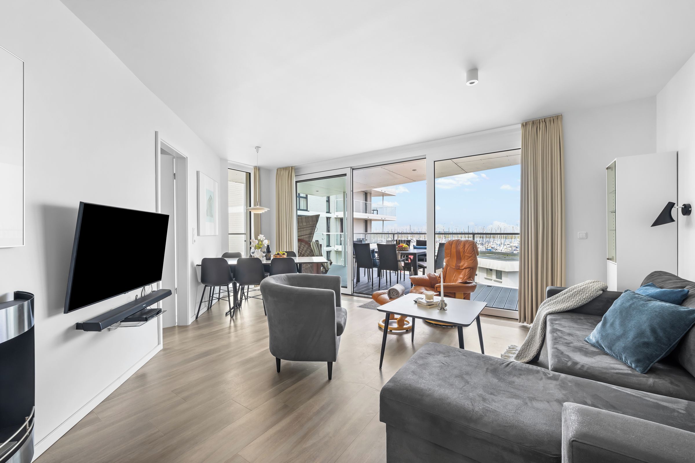 A modern living room with a flat-screen TV, a sofa, a dining area, and a balcony with a view of the city.
