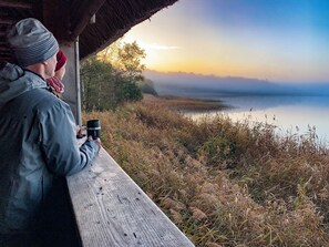 A person sitting at a wooden bench overlooking a calm body of water with trees and a clear sky.
