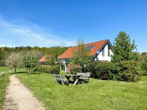 Landelijk tafereel met een grindpad dat leidt naar een houten bank en picknicktafel, omgeven door weelderig groen en een wit gebouw met een rood dak.