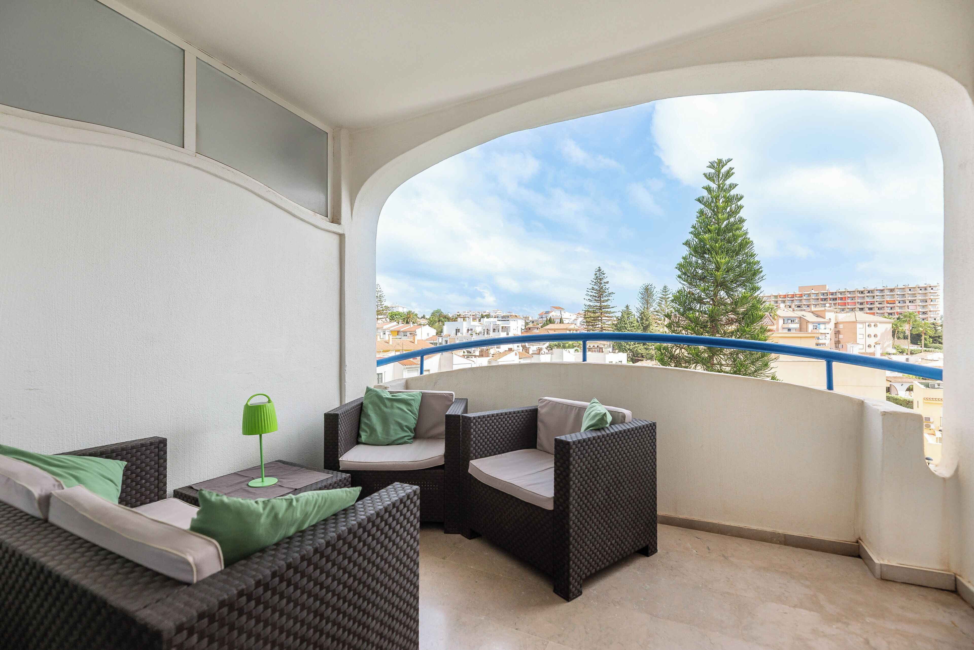 A balcony with wicker furniture, a table with a lamp, and a view of buildings and trees.