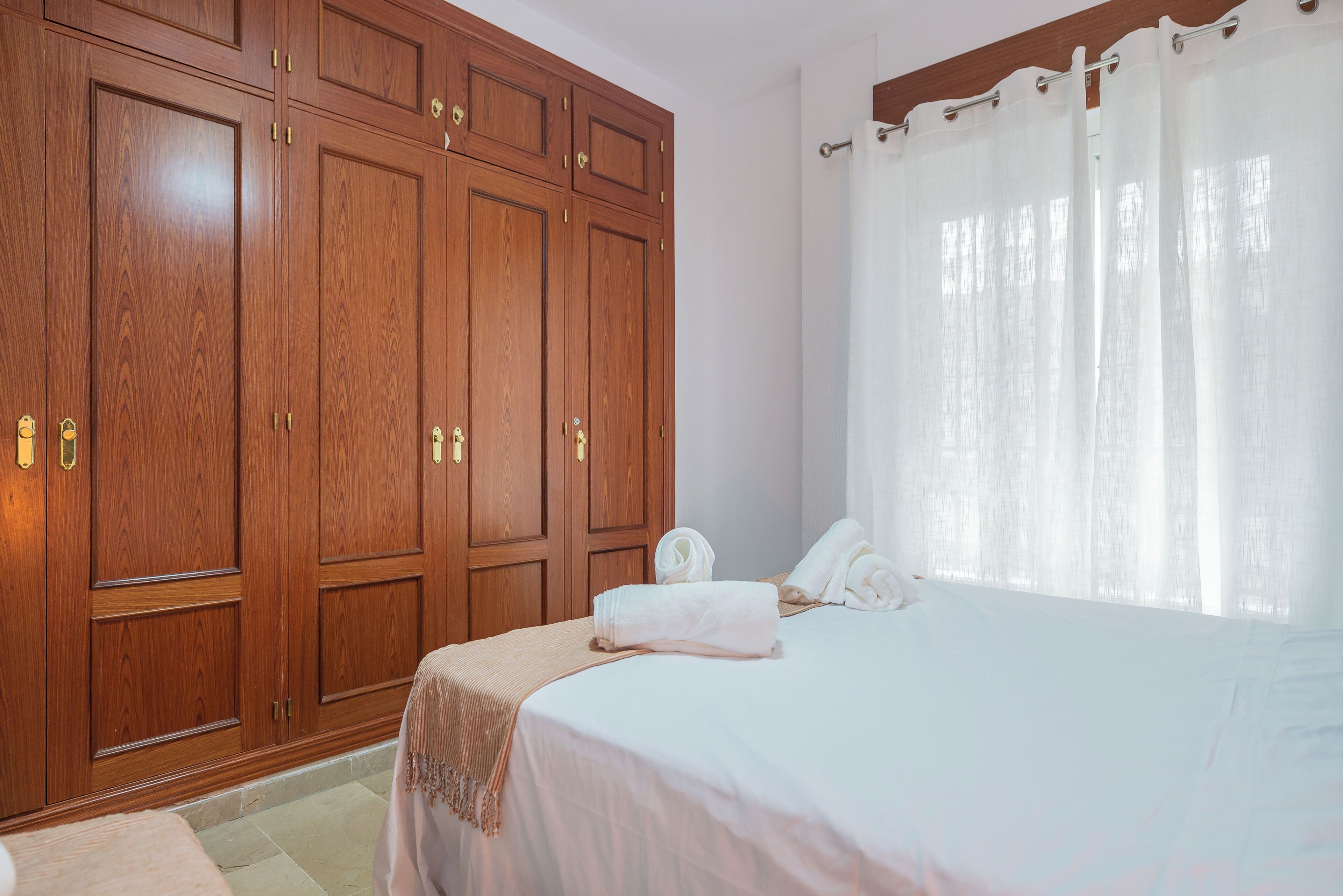 A bedroom with a large wooden wardrobe, a bed with white linens, and a window with sheer curtains.