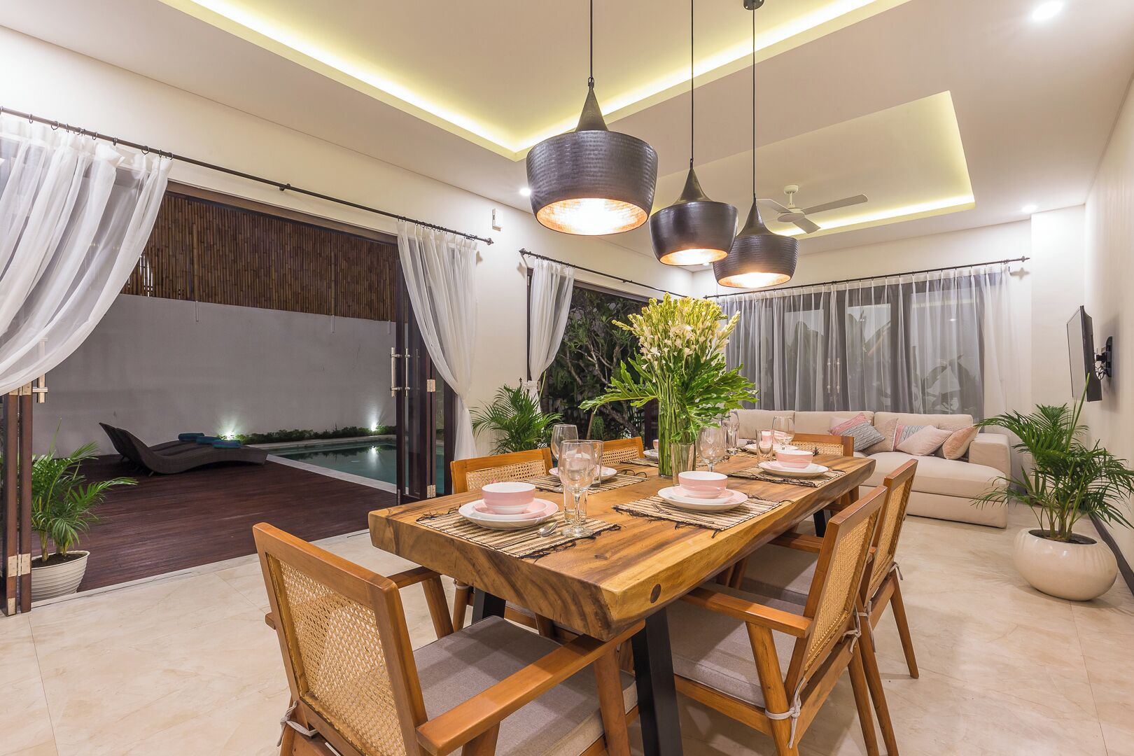 A dining area with a wooden table set for four, surrounded by chairs, with a view of a pool and a sofa in the background.