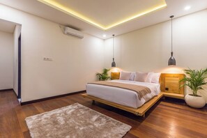 A modern bedroom with a wooden bed, bedside tables, a large potted plant, and a ceiling light fixture. - Villa Sereine Seminyak (Seminyak)