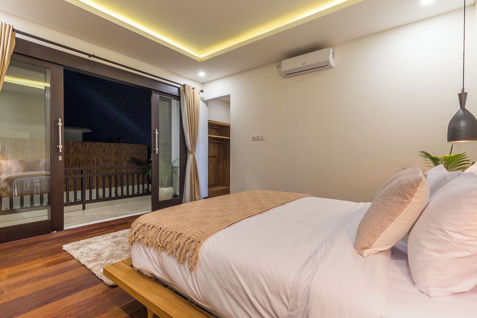 A bedroom with a bed, a bedside lamp, a sliding glass door leading to a balcony, and an air conditioning unit on the wall.