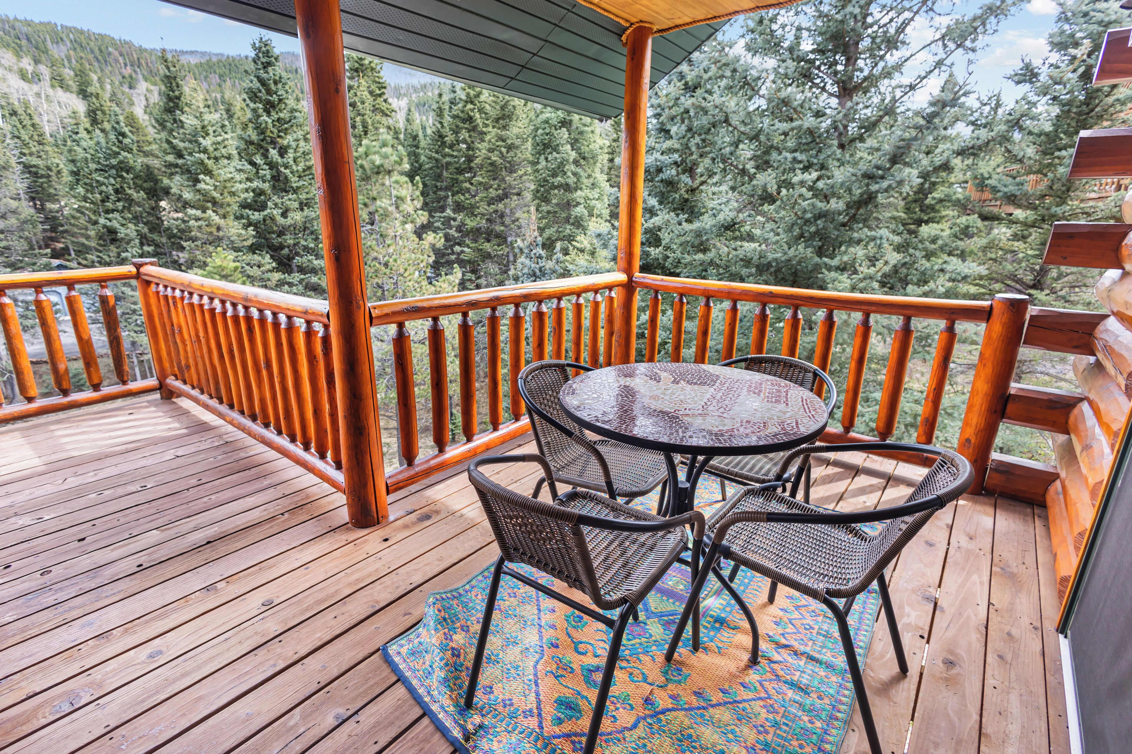 A wooden deck with a round table and four chairs, surrounded by a wooden railing, overlooking a forested area.