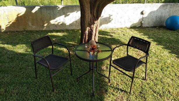 Een klein terras buiten met twee stoelen en een tafel onder een boom, omringd door een gazon en een witte muur.