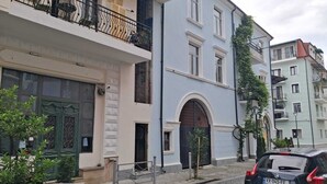 Calle con edificios de varios pisos, un coche estacionado al costado y un árbol pequeño en primer plano.