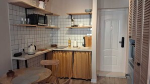 Cocina compacta con baldosas blancas, estantes de madera, un microondas, un hervidor y un comedor con mesa y sillas.