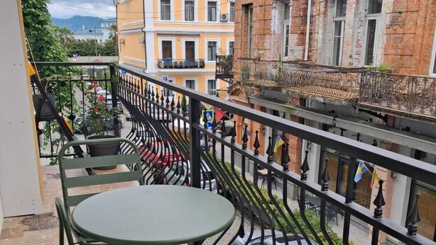Un balcón con una mesa y sillas, barandilla de metal y edificios al fondo.
