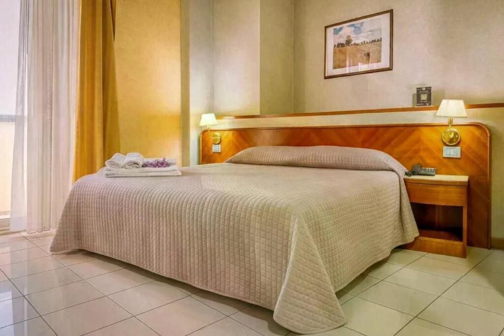 A hotel room with a bed, a nightstand, a lamp, a framed picture on the wall, and a window with curtains.