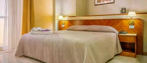 A hotel room with a bed, a nightstand, a lamp, a framed picture on the wall, and a window with curtains.