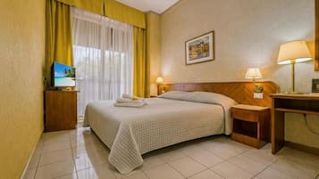 Una habitación de hotel con cama, escritorio, silla, televisor y ventana con cortinas.