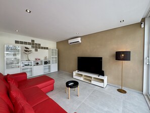 Ein modernes Wohnzimmer mit einem roten Sofa, einem Fernseher auf einem weißen Schrank, einem kleinen schwarzen Tisch und einer Stehlampe.