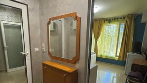 Ein Zimmer mit einem Holzkasten, einem Spiegel, einer Tür und Blick auf ein weiteres Zimmer durch eine geöffnete Tür.