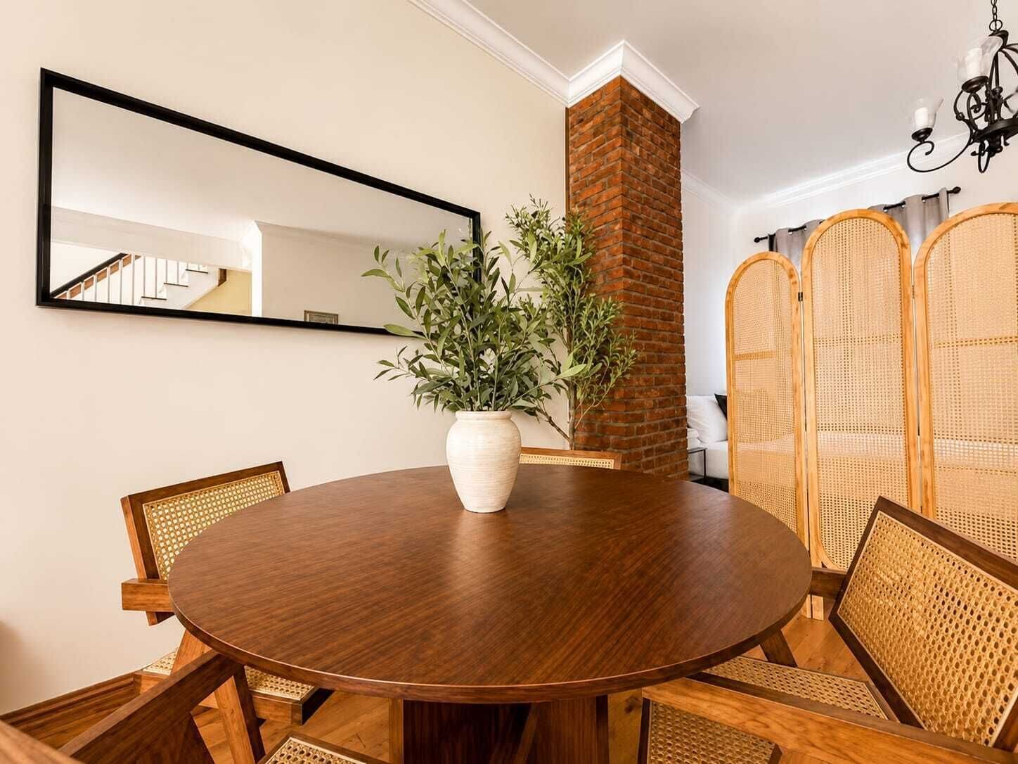 A round wooden dining table with a white vase, wicker chairs, a brick wall, a mirror, and a rattan partition.