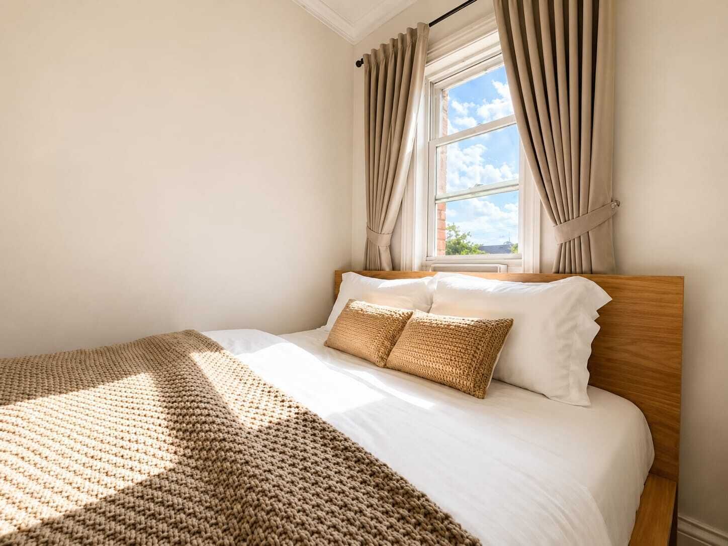 A neatly made bed with a wooden headboard, a window with curtains, and a view of the sky.