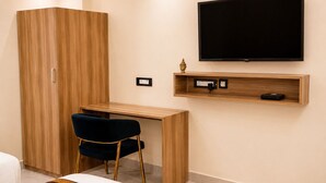 A bedroom with a bed, a desk with a chair, a wardrobe, a TV mounted on the wall, and a shelf with a statue. - Royal  Chandra Hotels (Patna)