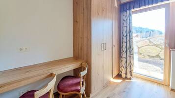 A room with a wooden desk, two chairs, a wardrobe, and a sliding glass door leading to an outdoor area with a stone wall.