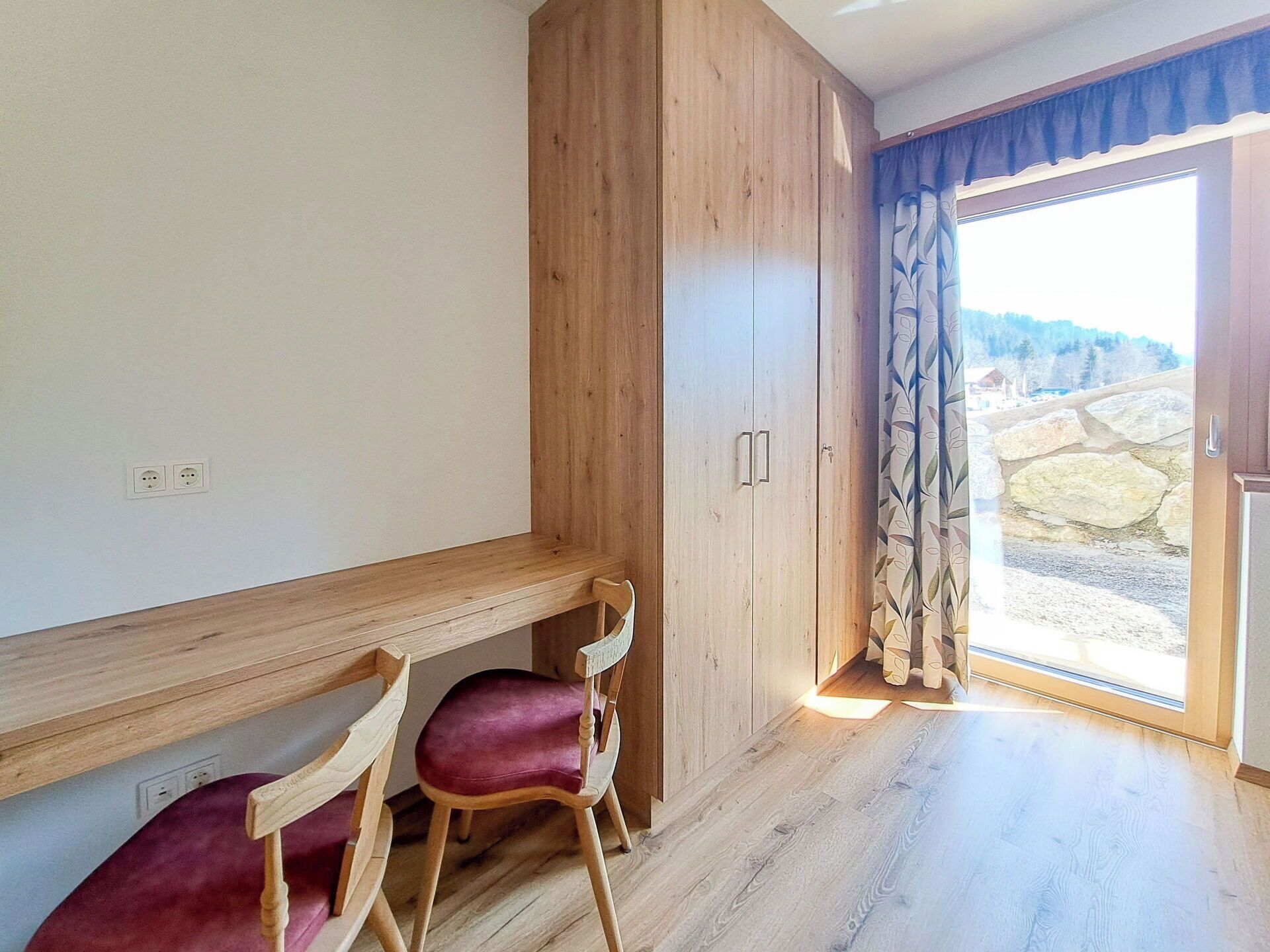 Una habitación con un escritorio de madera, dos sillas, un armario y un ventanal corredero que da a un espacio exterior con un muro de piedra.