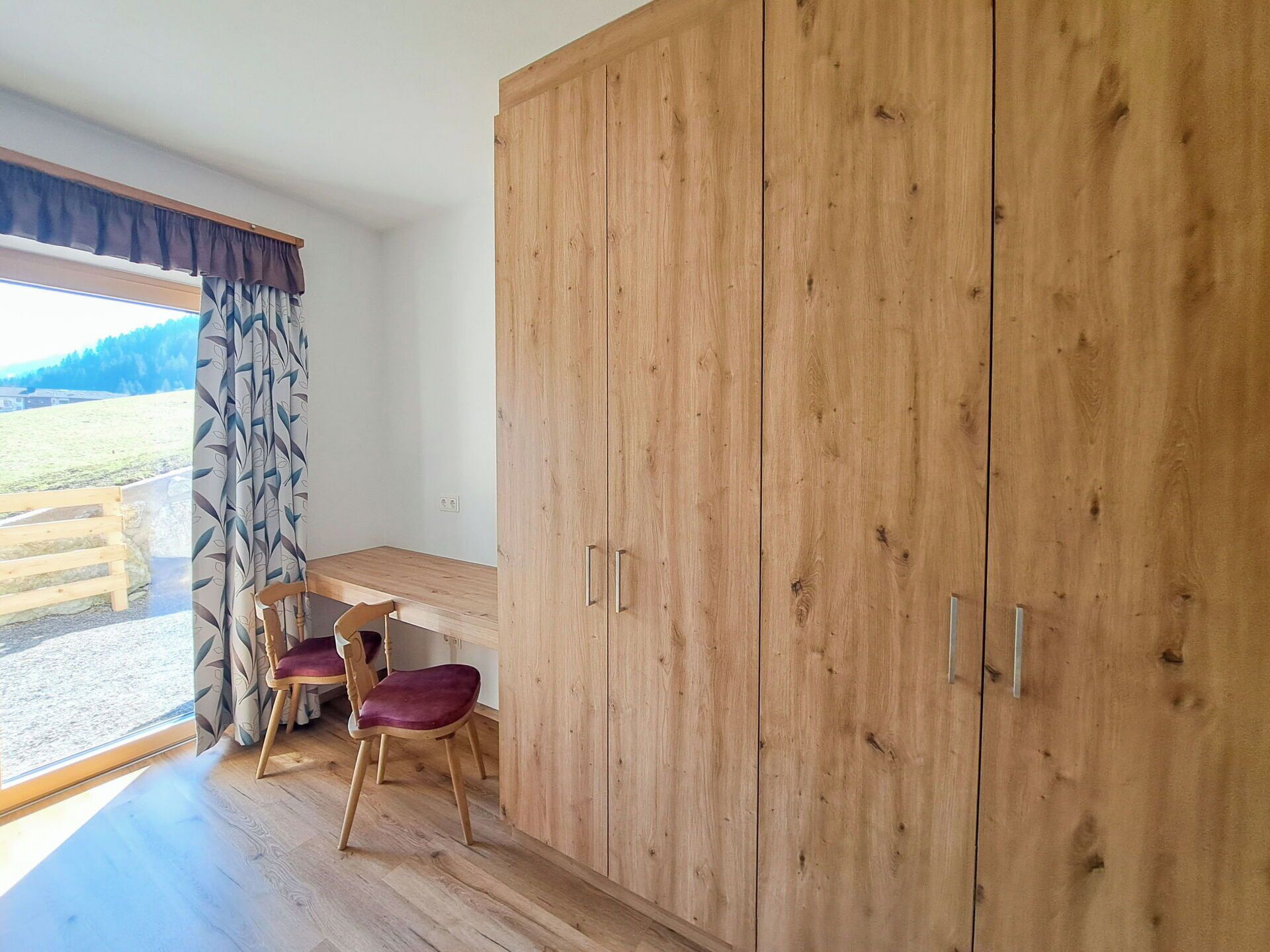 Una habitación con un armario de madera, un escritorio, dos sillas y una ventana con cortinas.