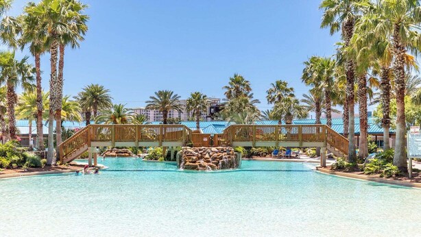 Ein Resort-Pool mit einer Holzbrücke, Palmen und blauem Himmel.