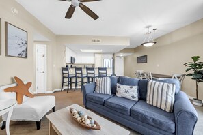 A living room with a sofa, a coffee table with decorative items, a ceiling fan, and a dining area with chairs and a table. - Sun, Sand & Gulf Views | End of Summer 15% off (Destin)