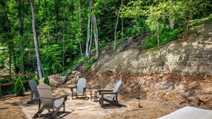 An outdoor seating area with Adirondack chairs and a table, surrounded by a forested hillside. - Tennessee Sunrise | Mountain Views, 3 King Suites, WifFi & Games (Sevierville)